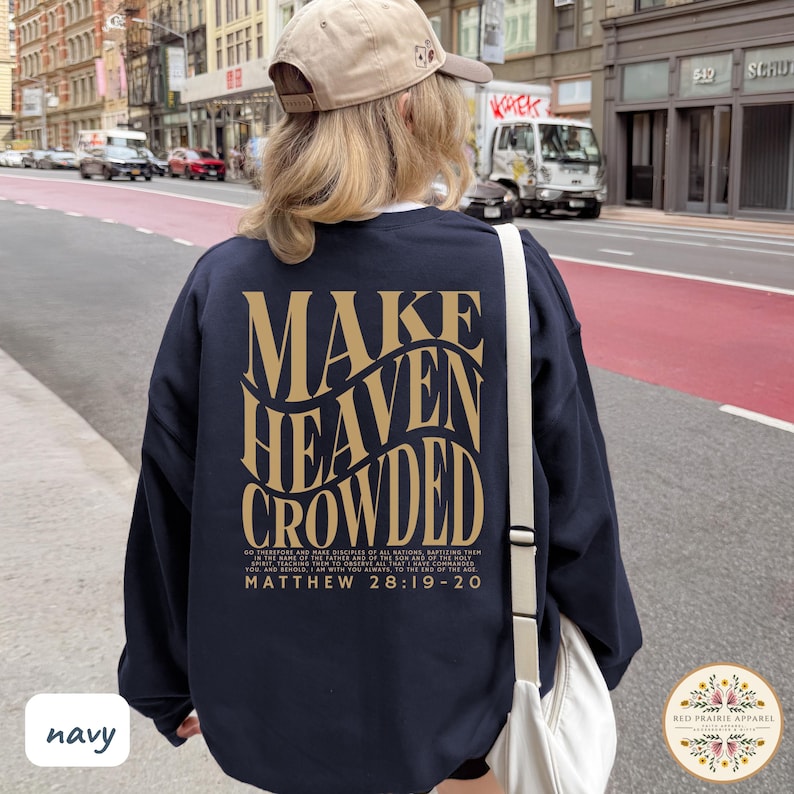 May include: A navy blue long-sleeve sweatshirt featuring the gold text "MAKE HEAVEN CROWDED" in a wavy font on the back. Below the main text, smaller gold text reads "MATTHEW 28:19-20" along with a passage from the Bible. A beige baseball cap is worn.