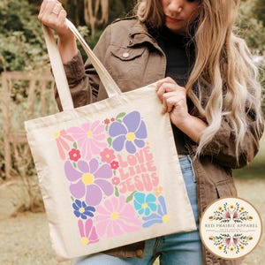 May include: A white canvas tote bag with a colorful floral design and the text "Love Like Jesus" in a retro font.