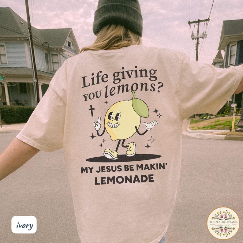 May include: Ivory-colored t-shirt with the text "Life giving you lemons?" and "MY JESUS BE MAKIN' LEMONADE." The graphic features a cartoon lemon character with a cross and stars. The shirt is a casual wear item.