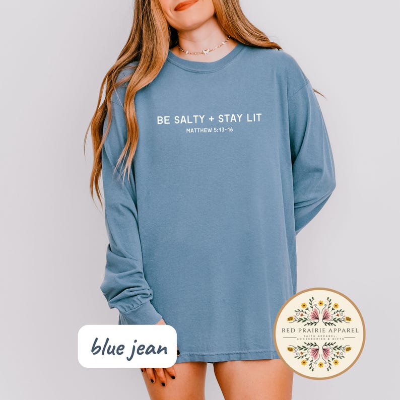 K&ouml;nnte beinhalten: Ein lang&auml;rmliges Shirt aus Jeansblau mit dem Text "BE SALTY + STAY LIT" und "MATTHEW 5:13-16" in wei&szlig;er Schrift.