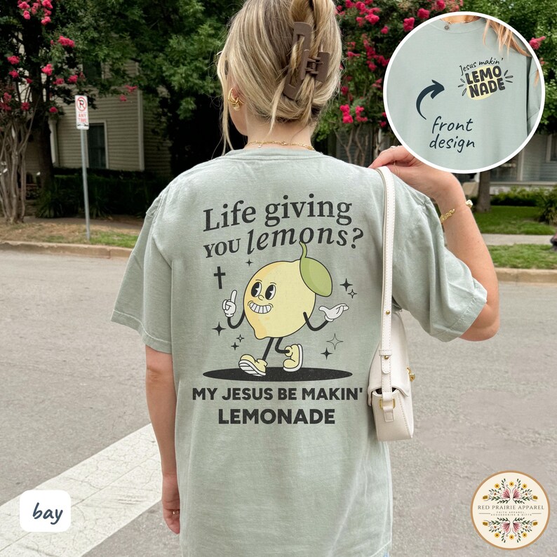 May include: Sage green t-shirt with a cartoon lemon character and the text "Life giving you lemons? My Jesus be makin' lemonade." The front design says "Jesus makin' lemonade."