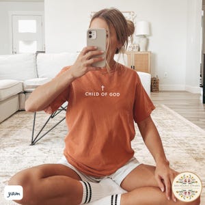 May include: Burnt orange t-shirt with the text "CHILD OF GOD" and a cross graphic. The shirt is a solid color and has a crew neck. The person is wearing white shorts and white socks with black stripes.