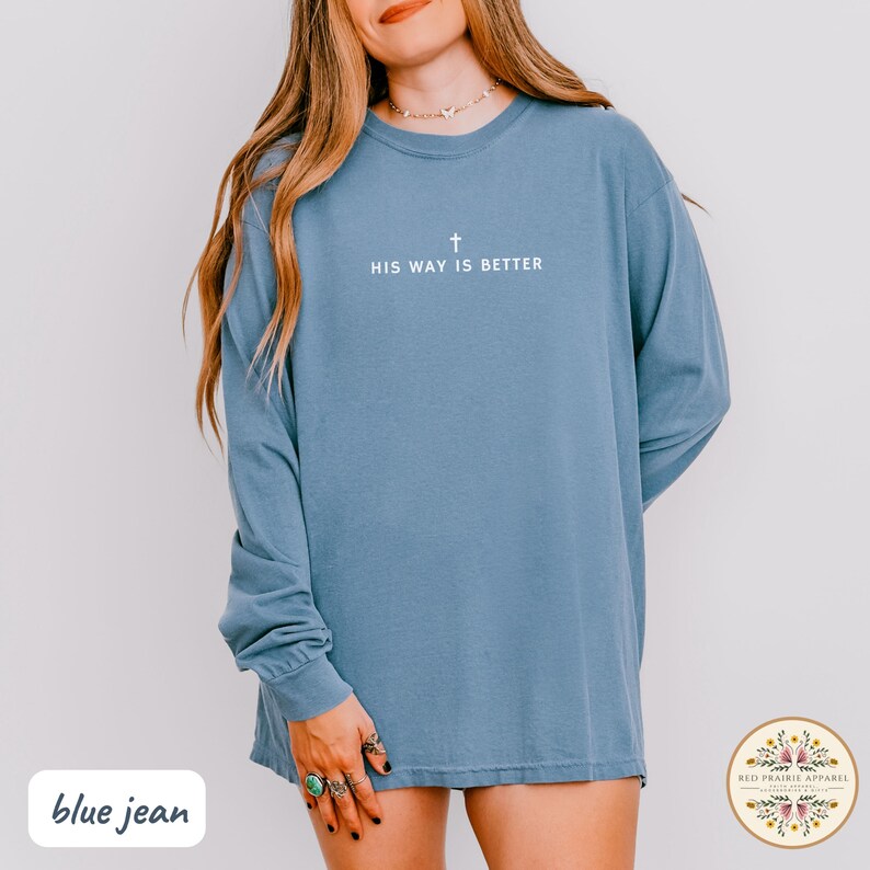 May include: A blue jean long-sleeve shirt with the text "HIS WAY IS BETTER" and a cross graphic. The shirt is a solid color and has a relaxed fit. The Red Prairie Apparel logo is in the bottom right corner.