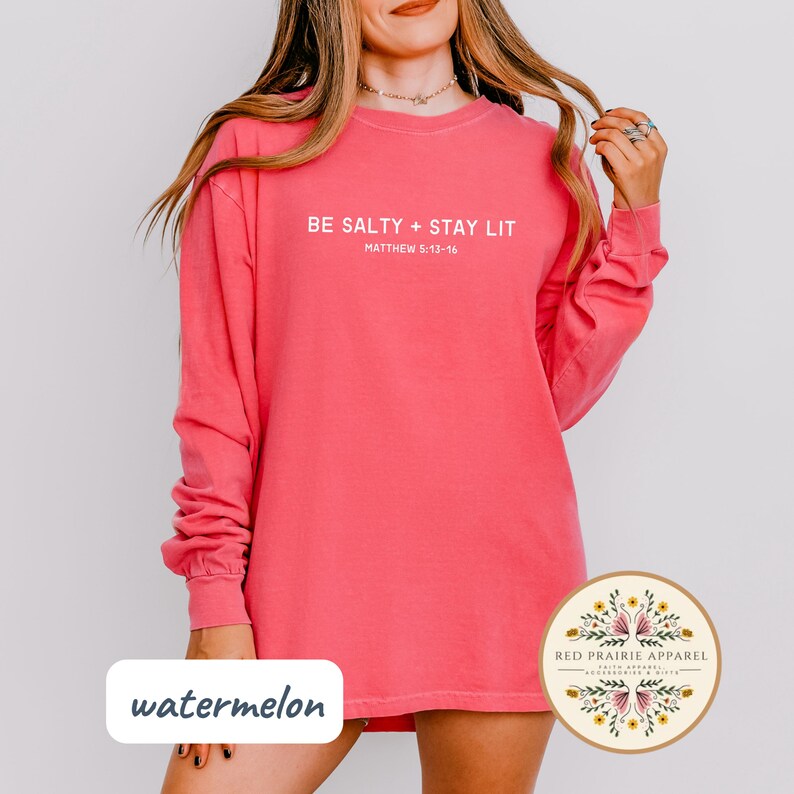 K&ouml;nnte beinhalten: Ein pinkfarbenes Langarmshirt mit dem Text "BE SALTY + STAY LIT" und "MATTHEW 5:13-16" in Wei&szlig;. Das Shirt ist aus einem weichen, bequemen Material.