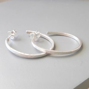 Könnte beinhalten: Ein Paar silberne Creolen. Die Ohrringe haben eine gehämmerten Textur auf der Außenseite und eine glatte, flache Innenseite. Die Stifte und Verschlüsse sind sichtbar. Die Ohrringe werden auf einer grauen Oberfläche präsentiert.