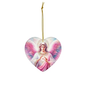 Puede incluir: Un adorno de cerámica en forma de corazón con una pintura de un ángel con alas rosadas sosteniendo un corazón en sus manos. El ángel está rodeado por un halo y un fondo suave rosa y azul.