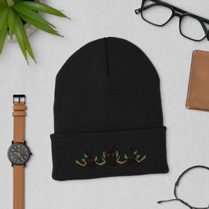 Puede incluir: Un gorro negro con un diseño bordado de tres insectos estilizados con ojos rojos y detalles verdes y dorados. El gorro está hecho de un material de punto suave, adecuado para el clima frío.