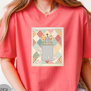 May include: A coral-colored short-sleeve t-shirt featuring a central rectangular graphic. The graphic displays a church building with a cross on top, set against a background of multicolored diamond-shaped quilt patterns in various floral prints.