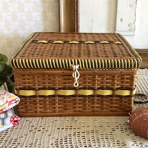 May include: A rectangular brown wicker sewing basket with a hinged lid, yellow accents, and a metal clasp. A stack of fabric squares, a pincushion, and scissors are nearby. The basket is approximately 10 inches wide.
