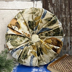 May include: A round, beige pillow with a floral and bird pattern. The pillow has a button in the center and is surrounded by folded fabric. A vintage book and a sprig of rosemary are also visible.