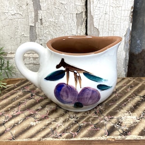 May include: A small, white ceramic creamer with a brown interior and a handle. The creamer is decorated with a painted design of purple plums and green leaves. The creamer is sitting on a brown and tan surface.