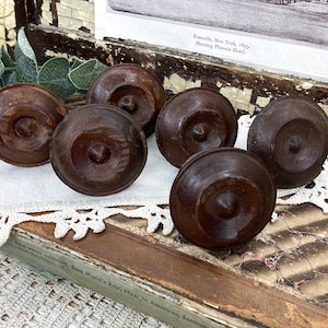 May include: Six antique, dark brown wooden knobs with a carved design. The knobs are arranged on a vintage book with a white lace doily. The knobs have a circular shape with a central, raised detail.