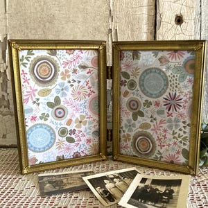May include: Two gold-framed photo frames with floral patterned inserts, displayed on a patterned surface. The frames are hinged together. Several vintage black and white photographs are in front of the frames. The background is a distressed white wooden surface.