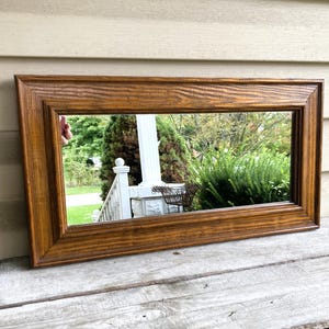 May include: A rectangular wall mirror featuring a medium-toned brown wooden frame with a visible grain pattern. The frame has a stepped, tiered profile that adds depth to the border. The reflective glass surface shows a clear outdoor reflection.