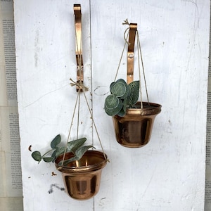 May include: Two copper-colored metal wall-mounted planters with curved brackets. Each planter is suspended by thin strings and contains a small green artificial plant with rounded leaves. The planters have a polished finish and a flared rim.