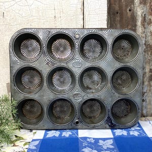 May include: Vintage 12-cup muffin tin with a silver finish. The tin has a rectangular shape with rounded cups, each featuring a radial pattern. The metal shows signs of wear, adding to its antique charm. A classic baking tool.