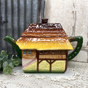 May include: A ceramic teapot shaped like a cottage with a brown thatched roof, yellow siding, and a green handle and spout. The structure features painted details including windows, wooden beams, and a textured base, creating a house-like appearance.