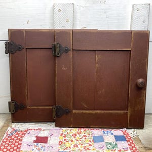 May include: Two rectangular cabinet doors with a matte reddish-brown finish and visible wear along the edges. Each door features dark metal decorative hinges and one small round wooden knob on the right side. The doors are positioned upright.