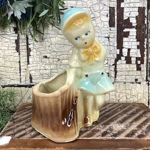May include: A vintage ceramic planter featuring a young child figure in a light blue dress and hat, with a yellow bow. The planter has a brown tree trunk design. The text on the book reads: First Premiums awarded at all Fairs where exhibited, from World's Fair, 1853, to American, Institute Fairs, inclusive, and at Philadelphia Centennial Exhibition, 1876.
