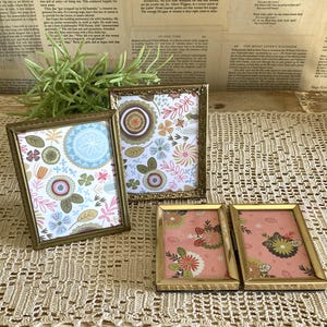 May include: Four rectangular picture frames with metallic gold-toned edges. Two frames are standing upright, displaying a white background with colorful floral patterns. Two frames are lying flat, featuring a pink background with floral designs.