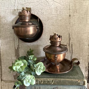 May include: Two copper oil lamps with rounded bowls, perforated burner collars, and wicks. One lamp is mounted on a circular wall plate, while the other sits on a flat base with a handle. A small green plant is placed in front of the lamps.