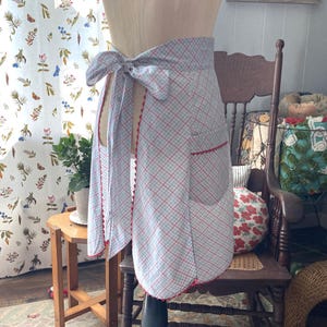 May include: A waist apron featuring a light blue and white plaid pattern with thin red diagonal lines. The apron has a rounded hem, a single patch pocket on the right side, and a tie closure at the back. Red scalloped trim edges the pocket and hem.