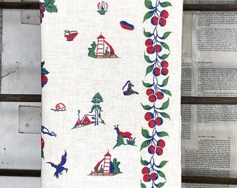 May include: A white kitchen towel with a repeating pattern of red, blue, and green illustrations of cabins, deer, and other outdoor scenes. A vertical border of red cherries and green leaves runs along the right side.