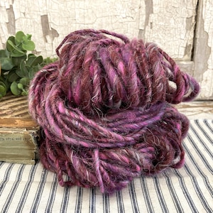 May include: A skein of thick, hand-dyed yarn in shades of burgundy, pink, and purple. The yarn is loosely spun, with a textured appearance. The background includes a wooden object, greenery, and a striped fabric.