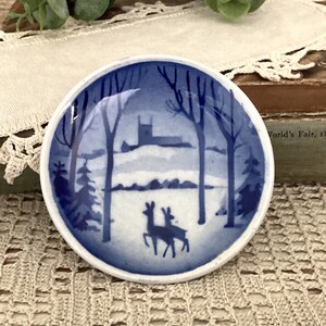 May include: A small, round ceramic dish featuring a blue and white transferware-style illustration of a winter landscape. The scene depicts two deer standing in a snowy forest with bare trees and a church building visible in the background.