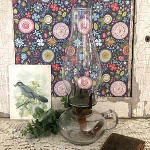 May include: A clear glass oil lamp with a rounded base, a handle, and a tall chimney. The lamp features a metal burner assembly with a visible wick. The design is functional, showcasing a transparent glass body that allows for a clear view of the internal wick structure and the overall construction of the lighting piece.