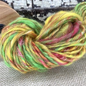 May include: A vibrant skein of hand-dyed yarn in shades of yellow, green, pink, and orange. The yarn is thick and textured, with a slight sheen. A wooden spool is visible in the background. Ideal for knitting or crochet projects.