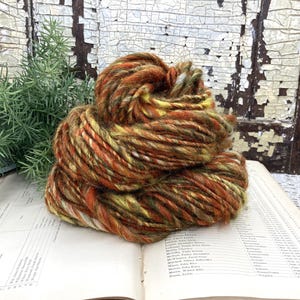 May include: A ball of hand-dyed yarn in shades of orange, yellow, and brown. The yarn is thick and textured, perfect for knitting or crocheting projects. The yarn is sitting on an open book with a weathered wooden background.
