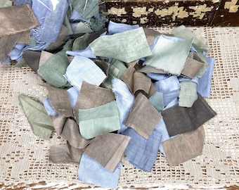 May include: A collection of fabric offcuts in shades of blue, green, and brown. The pieces are of irregular shapes and sizes, suggesting they are cut from different textiles. The scraps are arranged on a cream-coloured, open-weave surface.