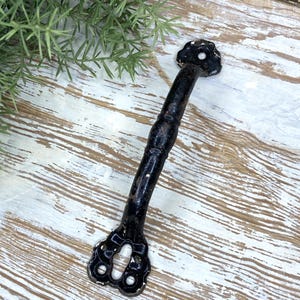 May include: A black metal door handle with a distressed finish and a cylindrical grip. The handle features two decorative, scalloped mounting plates at each end, each containing pre-drilled screw holes for attachment to a door or cabinet surface.