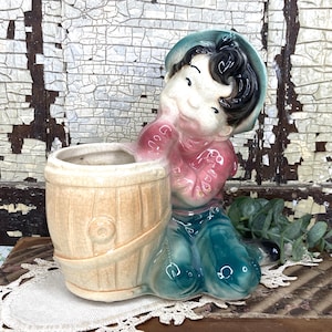 May include: A vintage ceramic planter in the shape of a boy holding a barrel. The planter features a boy in a blue hat, pink shirt, and blue pants. The barrel is beige with a light brown trim. The planter is likely from the mid-20th century.