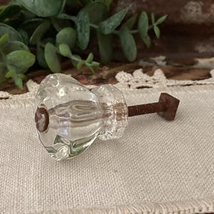 May include: A clear, faceted glass cabinet knob featuring a multi-sided, reflective surface. The knob is attached to a threaded, rusted metal bolt that extends outward, ending in a small, square-shaped metal base component.