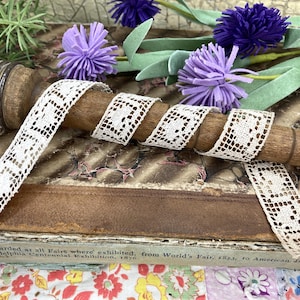 May include: Cream-colored lace ribbon with a geometric pattern is wrapped around a wooden spool. The spool rests on a vintage book with text. Purple pom-pom flowers and green foliage are in the background.