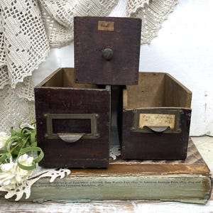 May include: Three dark brown wooden drawers with brass label holders and a small wooden knob. One drawer features a paper label with the text No. 6, while another displays a handwritten paper label. The drawers are stacked on a weathered book.