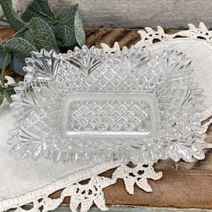 May include: A clear, rectangular glass dish with a diamond pattern and scalloped edges. The dish has a rectangular interior and is surrounded by a decorative border. The dish is sitting on a white cloth and a brown surface.