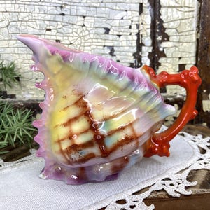 May include: A seashell-shaped creamer with a vibrant, iridescent finish. The creamer features a coral-like handle and spout, with colors ranging from pink and yellow to blue and brown. The creamer is a decorative piece.