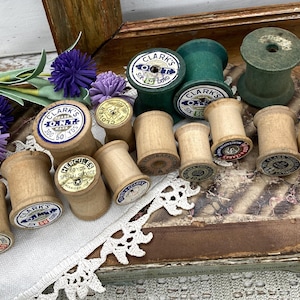 May include: A collection of vintage wooden spools with various labels, including "CLARK'S O.N.T." and "MERCERIZED 60 YDS SPECIAL SERVICE." Some spools are green, others are natural wood tones. The spools are arranged on a vintage book and lace.