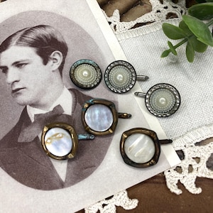 May include: An assortment of vintage cufflinks with pearl and mother-of-pearl insets. The cufflinks are in various shapes, including round and square, with a tarnished brass or bronze finish. A sepia-toned portrait of a man is in the background.