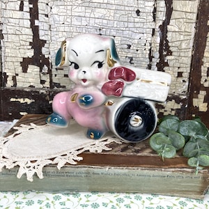 May include: A vintage ceramic figurine of a dog pulling a cart. The dog is white, pink, and blue with gold accents. The cart is white with red accents and a black wheel. The figurine is set on a book with text.