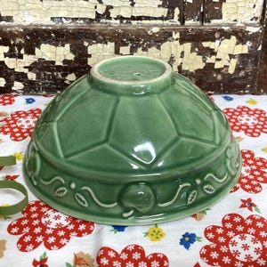 May include: A sage green ceramic bowl featuring a geometric faceted pattern on the upper half and a raised relief border along the base. The lower section displays a repeating motif of small apples and leaves connected by a continuous vine design.