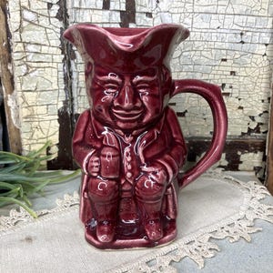 May include: A burgundy ceramic pitcher shaped like a man wearing a hat and holding a mug. The pitcher has a handle and a detailed face.