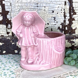 May include: A vintage pink ceramic planter featuring a young child under an umbrella. The planter has a textured, tree-trunk-like design with a small opening for plants. The child figure is in relief, adding depth to the piece.
