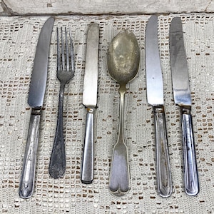 May include: A collection of six vintage silver-toned metal utensils arranged in a row, consisting of four dinner knives with decorative handles, one four-tined fork with a textured handle, and one large oval-shaped spoon with a smooth handle.