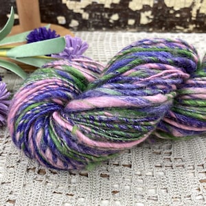 May include: A skein of hand-dyed yarn in shades of purple, pink, and green. The yarn is thick and textured, with a mix of colors that create a variegated effect. The yarn is likely for knitting or crochet projects.