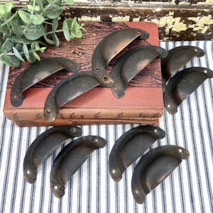 May include: A collection of antique, dark-colored metal drawer pulls arranged on a vintage book. The pulls have a curved, half-moon shape with two screw holes. The background includes greenery and a striped fabric.