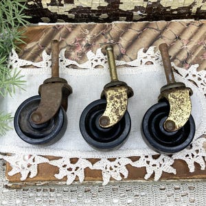 May include: Three vintage furniture casters featuring black wheels and weathered metal stems and brackets. Each caster has a central mounting post and a swiveling frame, showing signs of age with a tarnished, textured metallic finish.